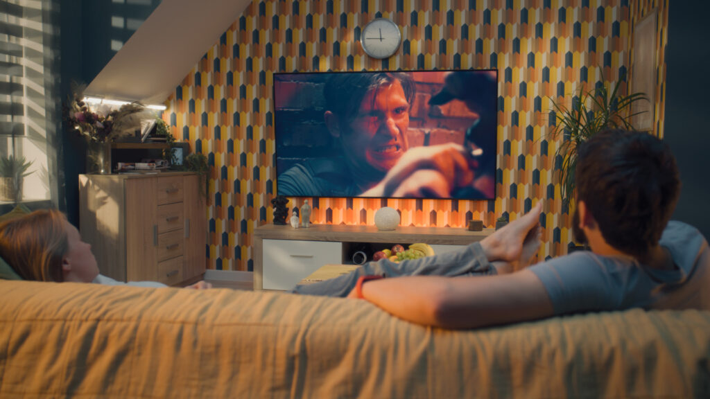Couple on sofa in living room, watching action movie on TV or criminal blockbuster on streaming service, talking and discussing acting, resting at home on weekend. Home theater in modern apartment.